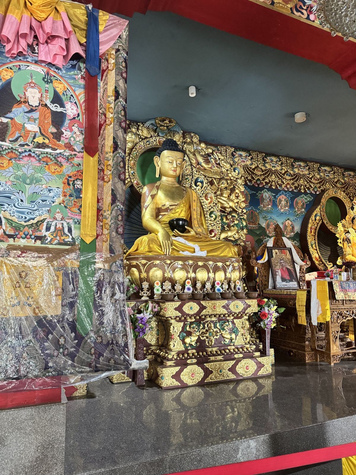 Golden Buddha statue on a carved lotus throne, surrounded by painted walls and a large thangka