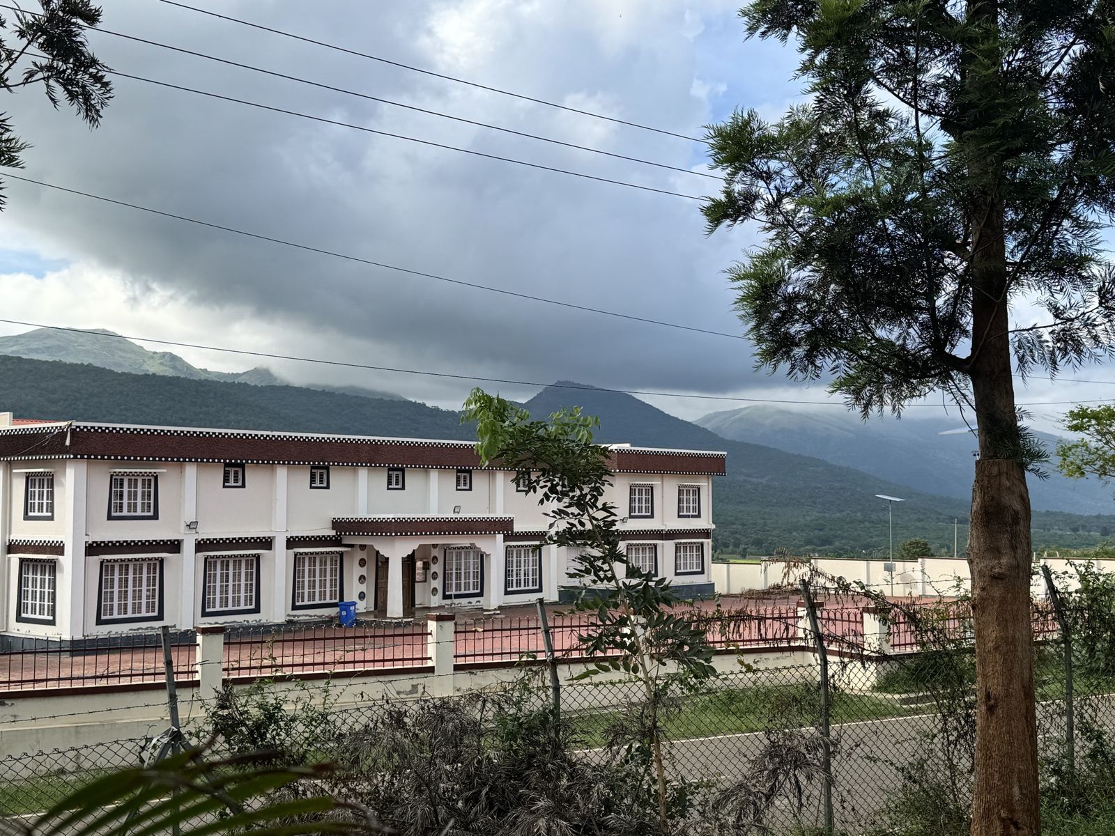A long white two-storey building with Tibetan-style maroon roof trim, with cloudy green hills behind it