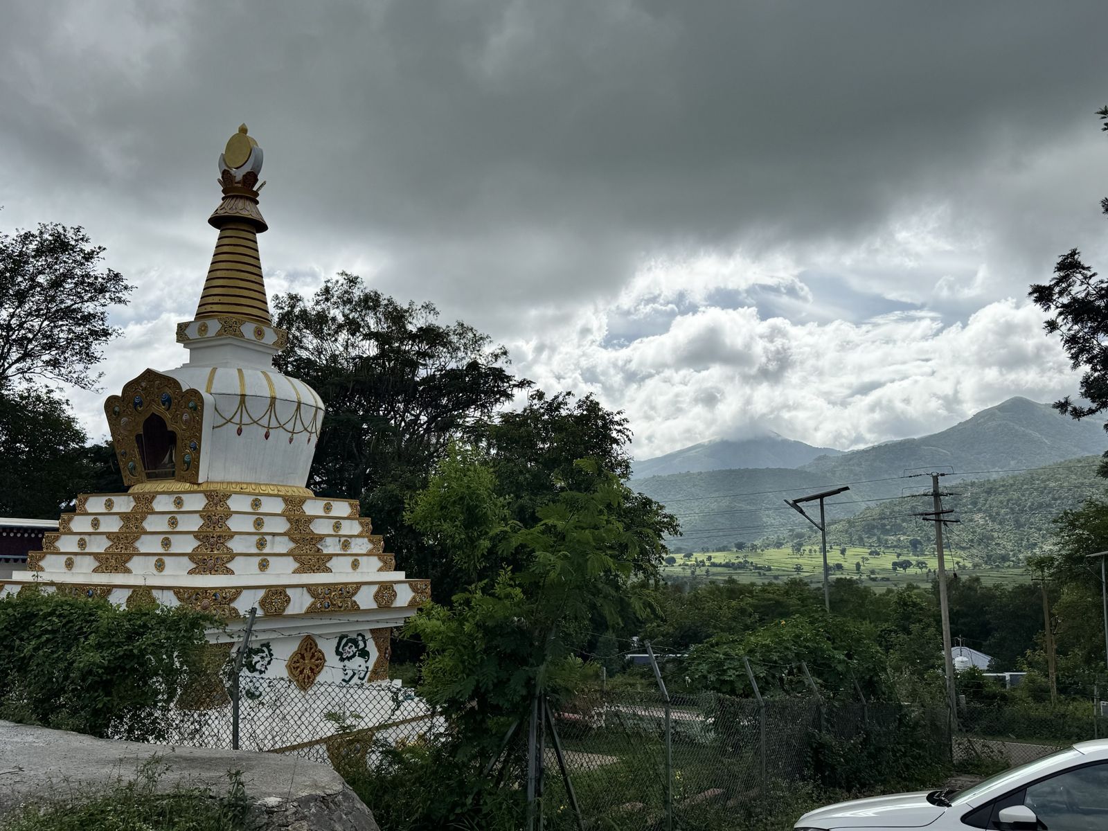 A single white and gold stupa on the left, with dark clouds over green hills and fields on the right