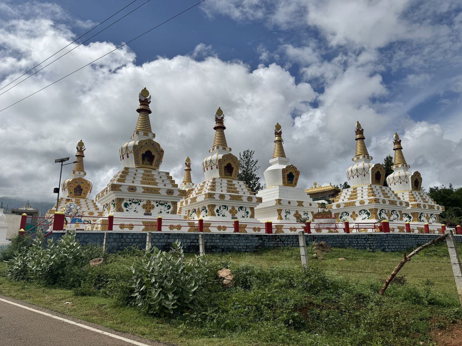 A row of white and gold stupas with red railings under a blue sky full of clouds